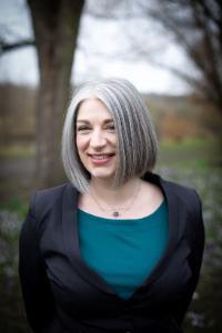 Sara Thornhurst, a white woman with short grey hair wearing a black jacket and green top. The background is blurred